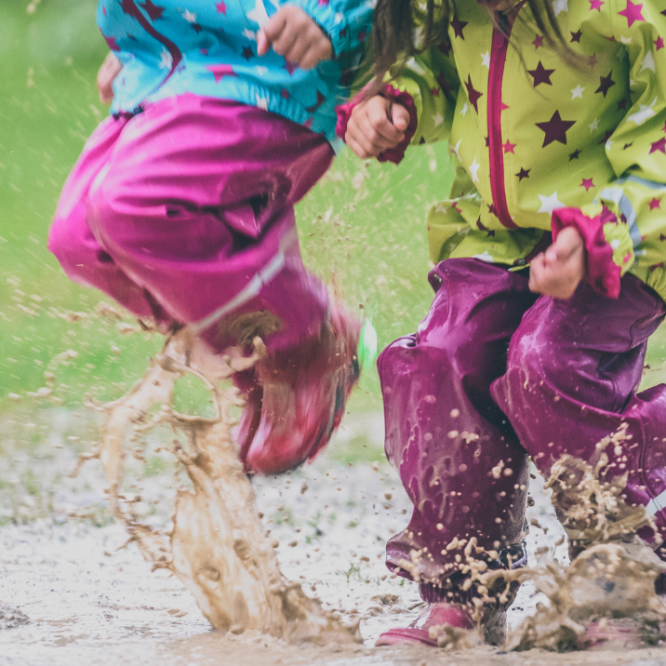 Two children jumping in muddy puddles in rain boots in spring